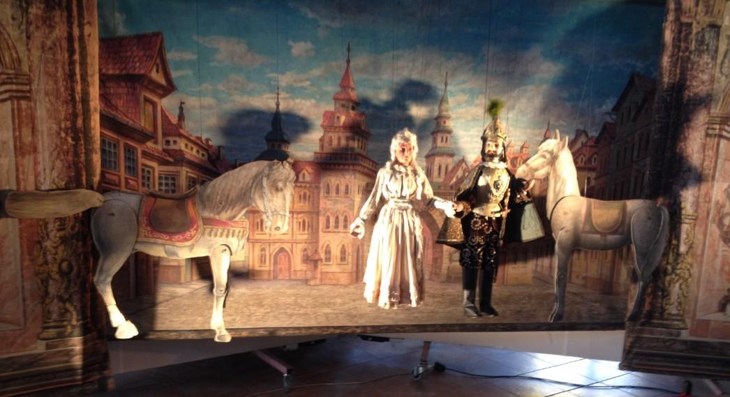 Detail loutkové scény s marionetami rytíře, dámy a koní na malovaném pozadí